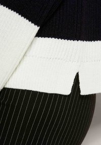 Gebreide trui met zwart-witte horizontale strepen. Heeft een geribbelde textuur en een zijsplit aan de zoom, gecombineerd met broek met fijne strepen.