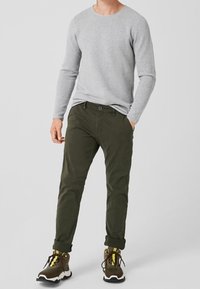 Chemise grise à manches longues, pantalon étroit olive, et baskets marron avec des accents jaunes, se tenant sur un fond neutre.
