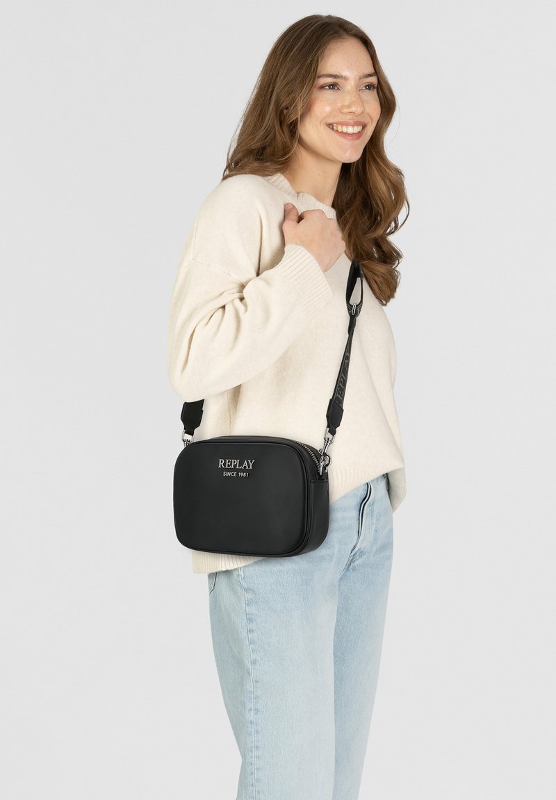 Bolso cruzado negro hecho de material suave, con forma rectangular, cierre con cremallera y logotipo impreso en la parte frontal. Correa ajustable.