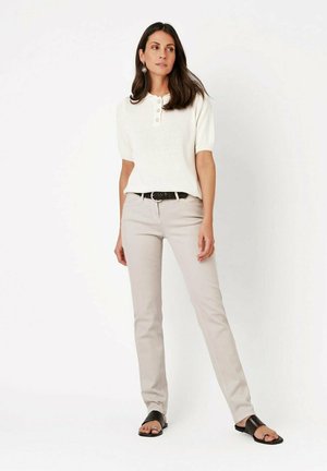 Frau steht mit entspannten Händen, trägt ein weißes kurzärmliges Strickoberteil, hellbeige Hose, schwarzen Gürtel und schwarze offene Sandalen auf weißem Hintergrund.