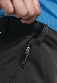 Schwarze Sportshorts haben eine glatte Textur, einen elastischen Bund und eine Reißverschlusstasche mit einem silbernen Ziehband. Ein blaues Shirt ist teilweise sichtbar.