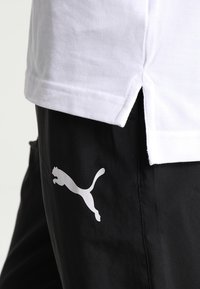 Bílá polo tričko s texturovaným materiálem, spárované s černými kalhotami, které mají velké bílé logo Puma na levé noze. Krátké postranní rozparky.