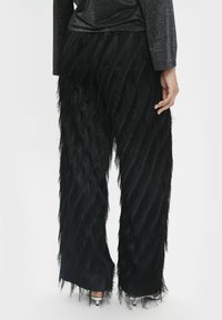 Schwarze Hose mit einem strukturierten Fransendesign, das ein wellenförmiges Muster und einen glänzenden Taillenbund aufweist. Hergestellt aus weichem Stoff mit lockerer Passform.