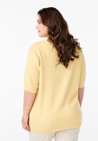 Gul kortærmet sweater lavet af blødt stof, med ribbede manchetter og en lidt længere bagkant, båret over lyse bukser.