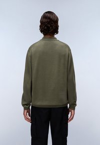 Olijfgroen pullover gemaakt van een zachte, gestructureerde stof. Voorzien van een geribde onderkant en manchetten, met een kraag en raglanmouwen. Zwarte cargobroek zichtbaar.