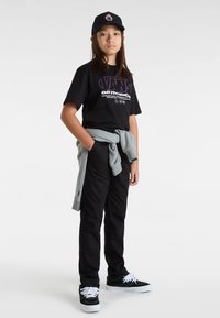Sort grafisk t-shirt med lilla tekst, grå sweatshirt bundet om taljen, sorte bukser og sorte sneakers med hvide snørebånd. Sort kasket med logo.
