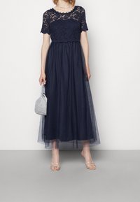 Mörkblå aftonklänning med spetstopp och korta ärmar, flödande tyllkjol, kompletterad med en glittrande handväska och sandaler med remmar.