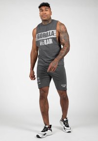 Grijze mouwloze shirt met de opdruk "GORILLA WEAR", gecombineerd met grijze shorts met logo's. Man draagt zwart-witte sport sneakers.