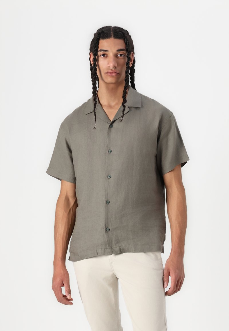 Camicetta in lino verde oliva a maniche corte con una vestibilità rilassata, dotata di colletto e bottoni leggeri, abbinata a pantaloni color crema.
