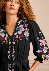 Robe noire à boutons, ornée de broderies florales en rose, bleu et vert. Elle présente une taille à nouer et des manches bouffantes en tissu texturé.
