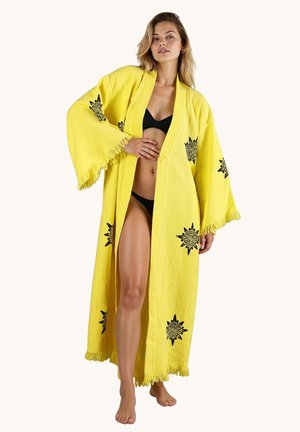 Kobieta ubrana w długi żółty szlafrok z czarnymi wzorami słońca na czarnym bikini, stoi boso na jednolitym tle.