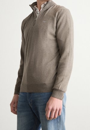 Homme portant un pull beige à fermeture éclair sur un chemisier blanc à col et un jean bleu, debout devant un fond uni.