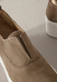 Des chaussures à enfiler en daim marron présentent un détail de couture à l'avant et une semelle en caoutchouc blanche. Le matériau a une apparence douce et texturée.