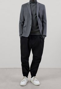 Blazer grigio sopra un dolcevita scuro, abbinato a pantaloni neri a sigaretta e sneakers bianche. Caratteristiche includono una tasca sul petto e polsini elastici.