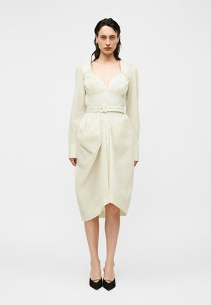 Kvinde med mørkt hår iført en off-white kjole med bælte og lange ærmer samt sorte spidse hæle, stående foran en ensfarvet hvid baggrund.