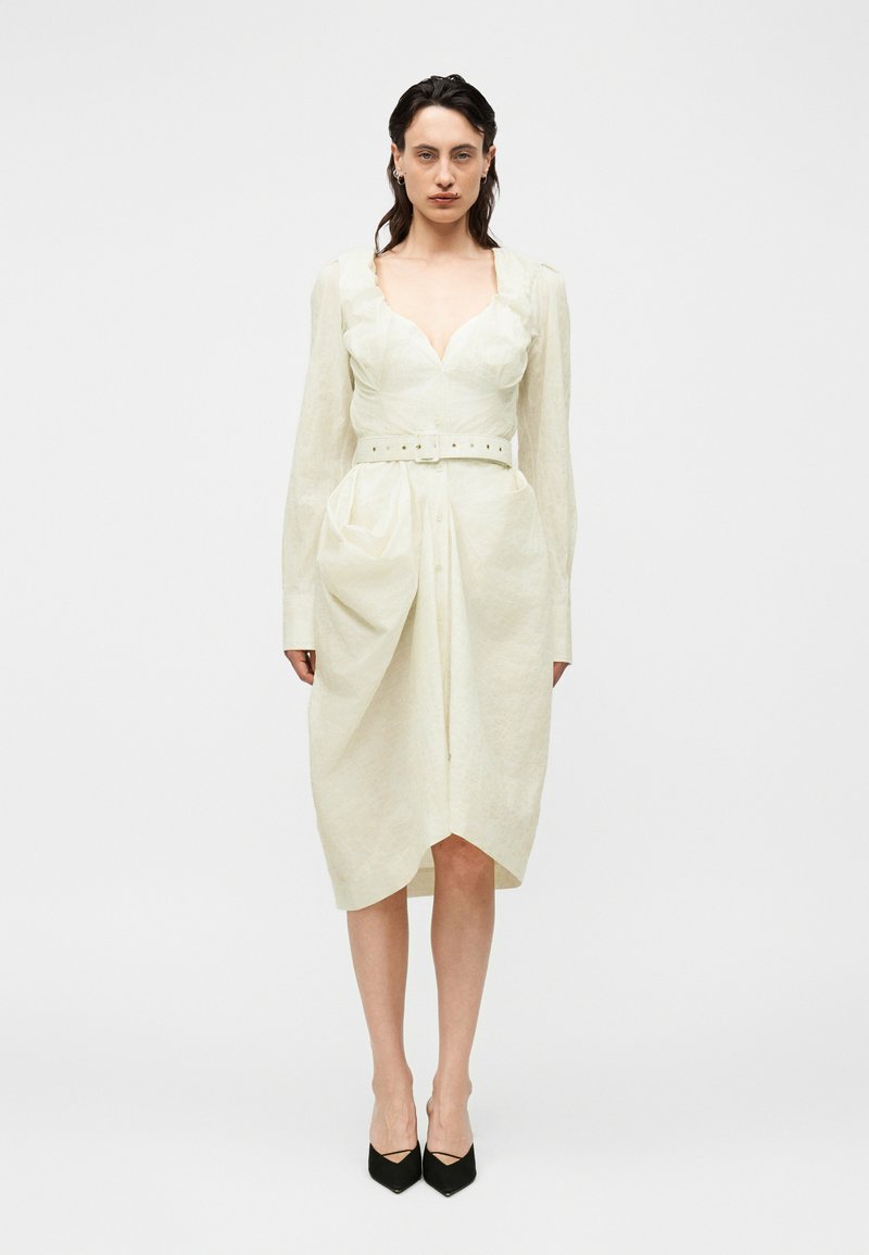 Femme aux cheveux foncés portant une robe ceinturée blanc cassé à manches longues et des escarpins noirs à bout pointu, debout devant un fond blanc uni.