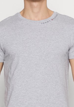 Mężczyzna noszący jasnoszary t-shirt z okrągłym dekoltem z małym logo Tommy Hilfiger blisko dekoltu na prawym ramieniu.