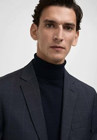 Navy checkered suit jacket with notched lapels, paired with a high-neck knit sweater in a matching dark hue, smooth texture and tailored fit.