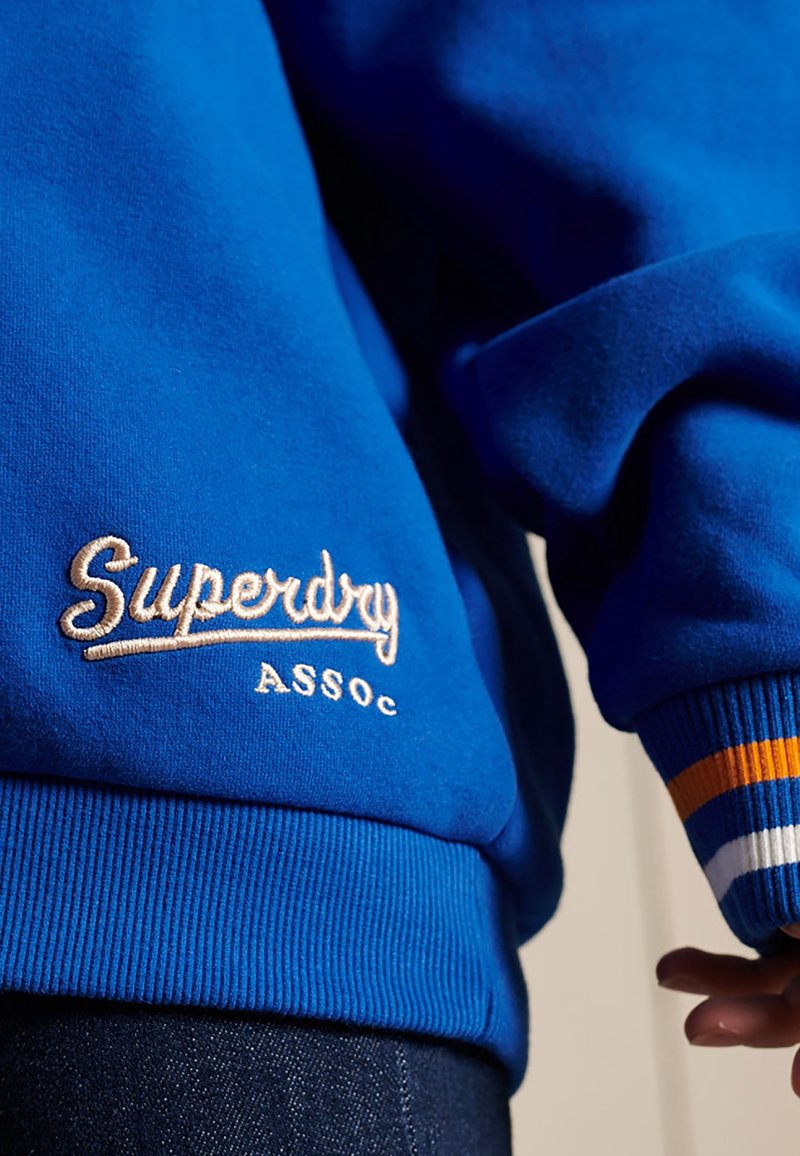 Sweat-shirt bleu avec le texte brodé "Superdry Assoc" et poignets côtelés avec des rayures blanches, orange et bleues, porté avec un jean foncé.