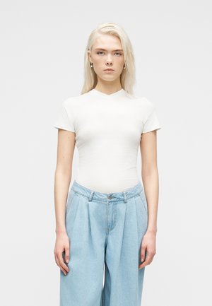 Blonde woman with neutral expression wearing fitted white short-sleeve top and high-waisted loose light blue jeans, standing against gray background.