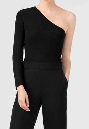 Woman wearing black ribbed one-shoulder top with long sleeve on left arm and black high-waisted pants, standing with right hand in pocket.