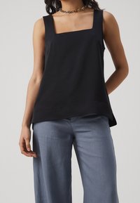 Top nero senza maniche con scollatura quadrata. Realizzato in tessuto liscio, presenta una vestibilità comoda e orlo dritto, abbinato a pantaloni azzurri chiari.