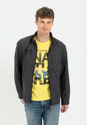 Black lightweight jacket with a zippered pocket, paired with a bright yellow T-shirt featuring bold black lettering, worn with blue jeans.