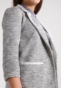 Femme portant un blazer gris texturé aux manches retroussées sur une chemise grise, visible de la taille au menton sur un fond uni.