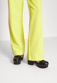 Svarta plattformsträskor med detaljer i guld, bärs med ljusgula vida byxor. Byxorna har en slät textur och en avsmalnande nederkant.