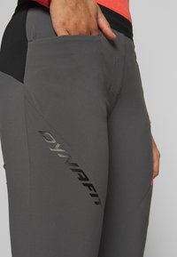 Šedé sportovní kalhoty s těsným střihem, s černým akcentem a boční kapsou. Na stehně černé logo "DYNAFIT".