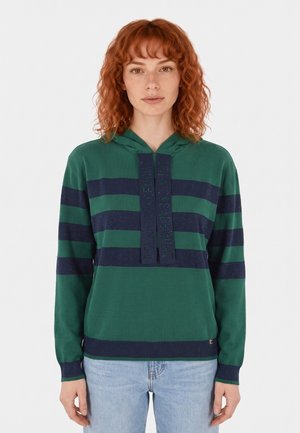 Donna con capelli rossi ricci che indossa una felpa con cappuccio a righe verde e blu navy e jeans azzurri, in piedi davanti a uno sfondo bianco uniforme.