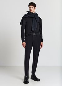 Schwarzes langärmeliges Hemd, enganliegende schwarze Hose und schwarze Chelsea-Stiefel. Ein dunkler, gerippter Schal ist lässig um den Hals gelegt.