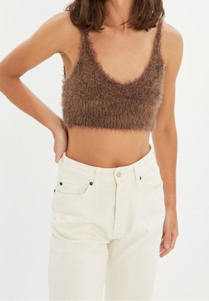 Frau trägt ein braunes, flauschiges, bauchfreies Top und weiß-hochgeschnittene Jeans, posiert mit einer Hand in der Hüfte vor weißem Hintergrund.