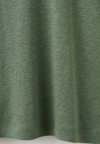 Tissu doux vert avec une texture subtile et des coutures visibles le long du bord inférieur, suspendu avec de légers plis.