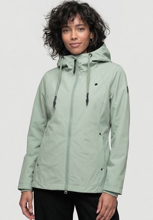 Woman with curly hair wearing a light green hooded jacket with zipper and drawstrings, standing against a plain gray background.