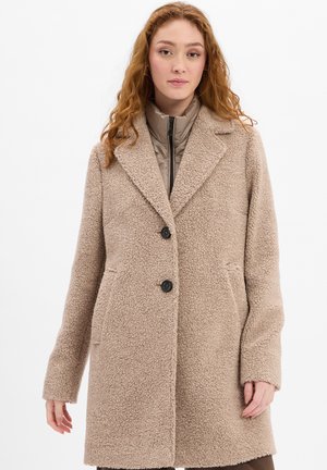Frau mit roten lockigen Haaren, die einen beigen Sherpa-Mantel über einer taupefarbenen Steppjacke trägt und vor einem weißen Hintergrund steht.