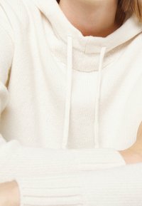 Jasnokremowa bluza z kapturem wykonana z miękkiej dzianiny, z wysokim kołnierzem, regulowanymi sznurkami i ściągaczami.