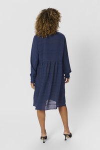 Abito blu navy a maniche lunghe con vita arricciata, tessuto testurizzato e pannelli trasparenti. Indossato con scarpe con tacco alto nere, di spalle.