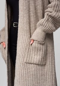 Beige Strickcardigan mit strukturiertem Stoff, gerippten Bündchen, zwei aufgesetzten Taschen und einem offenen Design. Weiche und oversized Passform.