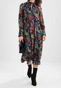 Robe maxi florale en tissu sombre avec un motif de feuilles vif en vert, rouge et bleu. Manches longues et détail de décolleté en forme de trou de clé.