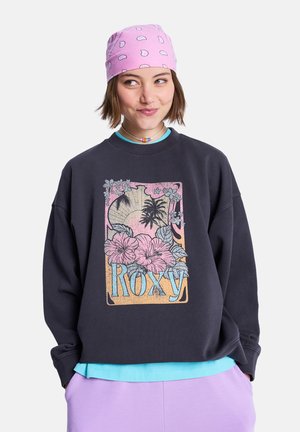 Mladý človek s krátkymi vlasmi, ktorý má na sebe ružovú bandanu a čiernu mikinu s tropickým kvetinovým vzorom a nápisom „Roxy“, ruky v kapsách levanduľových nohavíc.