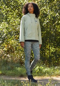Hellgrüne, Reißverschluss-Jacke mit großen Taschen und Kapuze; kombiniert mit grauen, tigerstreifen Leggings und schwarzen Stiefeletten. Natürlicher Hintergrund.
