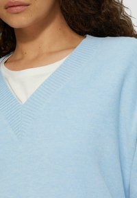 Lichtblauwe v-hals trui met ribbeltextuur bij de halslijn, gedragen over een witte ronde hals t-shirt. Zacht gebreid materiaal, casual ontwerp.