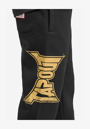 Mustad, pehmest materjalist dressipüksid, mille vasakul jalal on suur kuldne kulunud stiilis tekst "XAPOUL".