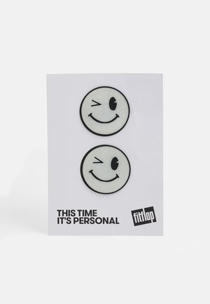 Dois patches de rostos sorridentes com contornos pretos em um cartão branco. Um pisca o olho; ambos têm uma textura brilhante. O texto diz "DESSA VEZ É PESSOAL."