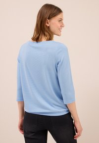 Frau mit hellbraunem Haar, die ein hellblaues Oberteil mit drei Viertelärmeln und schwarze Jeans trägt. Sie ist leicht nach rechts gedreht, vor einem neutralen Hintergrund.