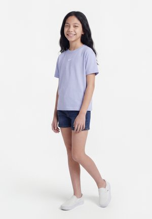 Fille souriante debout, portant un t-shirt Nike violet clair, un short en denim foncé et des baskets blanches sans lacets sur un fond blanc.