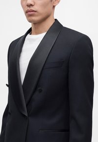 Homme portant un blazer noir croisé avec des revers en satin sur un t-shirt blanc à col rond, cadré sous les yeux.