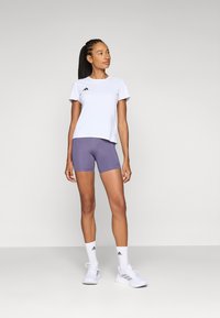 Hvid t-shirt med korte ærmer og sort logo, parret med lilla shorts og hvide træningssko, iført hvide ankelsokker.