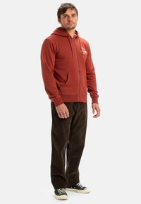 Sweat à capuche zippé rouge avec une poche frontale et un cordon de serrage, associé à un pantalon en velours côtelé sombre et des baskets noires.